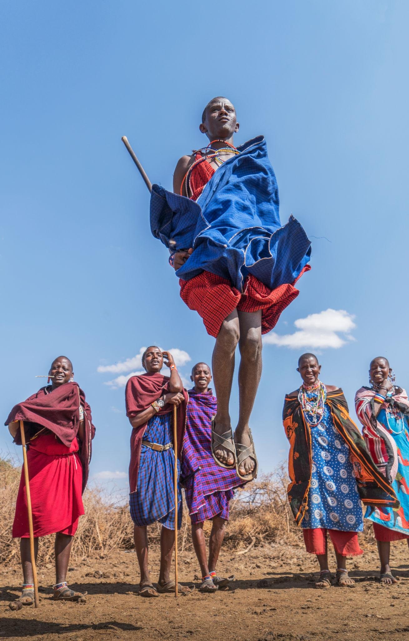 A vibrant and colorful portrait of a Maasai elder in traditional red robes and intricate beadwork. He is sharing a story with a small group of visitors under a large shade tree. The setting is warm and inviting, emphasizing the deep cultural connections and authentic human experiences offered during Tanzanian day trips and cultural immersions.