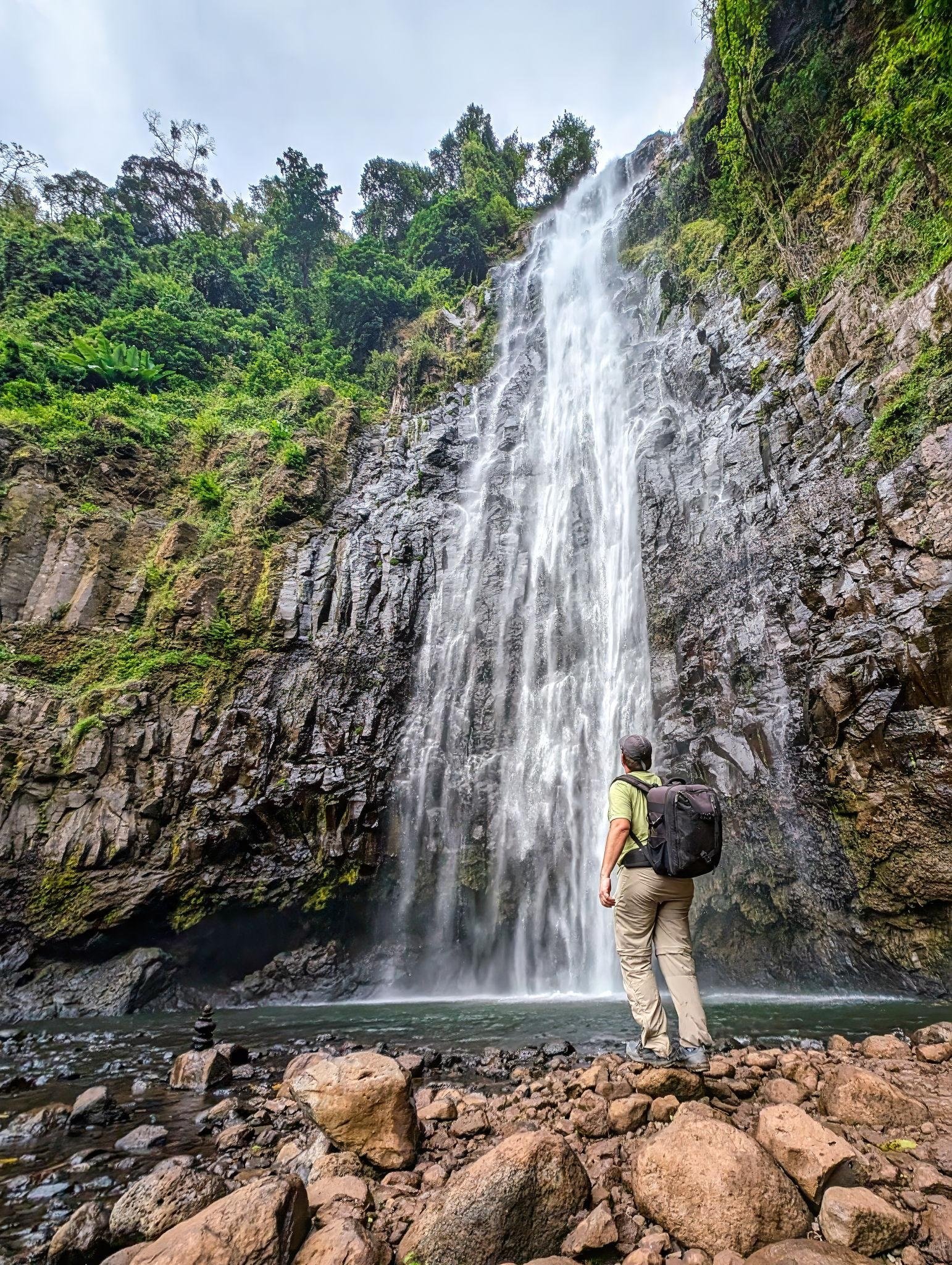 A crystal clear waterfall cascading down a lush green cliff in the Materuni region near Kilimanjaro. The surrounding vegetation is tropical and vibrant. A fine mist is visible in the air, caught by the sunlight. The mood is refreshing and peaceful, highlighting the hidden gems found on Tanzanian day trips.